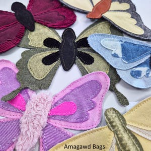 Op de afbeelding: Een verzameling stoffen vlinderapplicaties in diverse kleuren, waaronder bordeauxrood, olijfgroen, lichtblauw, roze en geel. De vlinders zijn voorzien van gestructureerde stiksels en gelaagde stoffen vleugels. De tekst op de afbeelding luidt Amagawd Bags by Miecaéla.