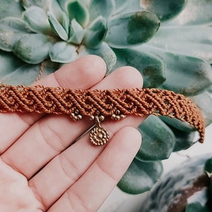 以下が含まれることがあります： ゴールドのチャームが付いたブラウンのミサンガアンクレット。アンクレットは編み紐で作られており、小さな丸いチャームがぶら下がっています。