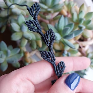 Pode incluir: Um suporte de plantas de macramé preto com um design delicado de folhas. O suporte é feito de corda de nó e apresenta uma série de pequenas folhas intrincadas.