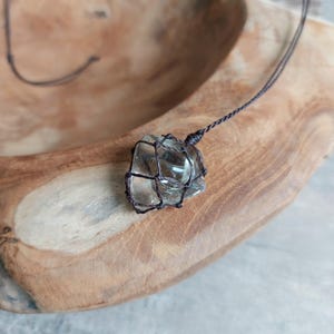 May include: A clear glass stone pendant wrapped in a brown macrame cord necklace. The necklace is displayed on a wooden surface.