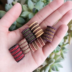 Könnte beinhalten: Fünf handgefertigte Makramee-Armbänder aus Kordel in verschiedenen Farben, darunter Rot, Braun, Grün und Gelb. Die Armbänder sind in einer Hand übereinander gestapelt.