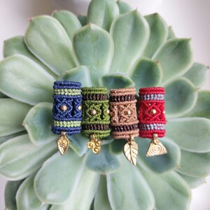 Op de afbeelding: Vier kleurrijke macramé haar kralen met goudkleurige bedels, rustend op een vetplant. De kralen zijn blauw, groen, bruin en rood, elk met een uniek ontwerp en bedel. De bedels zijn blad- en bloemvormen.