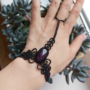 Puede incluir: Pulsera de mano de macramé negro con una piedra preciosa ovalada púrpura. La pulsera se conecta a un anillo con una delicada cadena. El diseño de macramé tiene intrincados nudos. La pulsera se lleva en una mano.