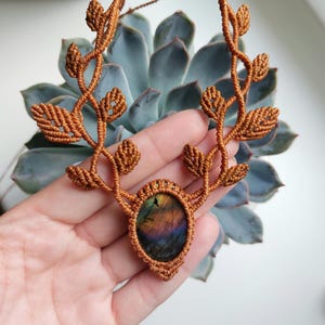 May include: A macrame necklace featuring a central oval labradorite gemstone with iridescent purple and gold flashes. The stone is encased in copper-colored cord, which extends upward into two symmetrical branches adorned with intricate leaf-shaped knots.