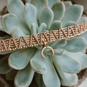 Peut inclure: Un bracelet tressé marron avec des perles dorées et un pendentif en forme de croissant de lune doré. Le bracelet est présenté sur une plante succulente verte.