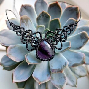 Può includere: Una collana in macramè nero con una pietra centrale a forma di goccia di colore viola e bianco. Il cordino è annodato in modo complesso con un motivo simmetrico che si estende dalla pietra, creando un design decorativo a forma di colletto.