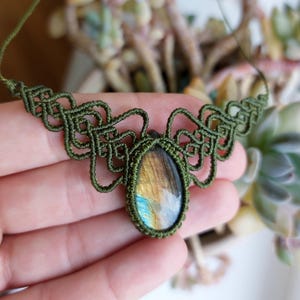 May include: A necklace featuring a teardrop-shaped labradorite gemstone with iridescent blue and gold flashes, encased in an intricate olive green macrame cord setting with symmetrical knotted patterns extending along the sides of the pendant.