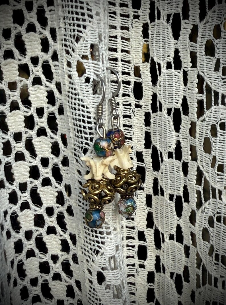 K&ouml;nnte beinhalten: Ein Paar Ohrringe mit einem knochenf&ouml;rmigen Anh&auml;nger und bunten Emailleperlen. Die Perlen sind blau, gr&uuml;n, rot und wei&szlig;.