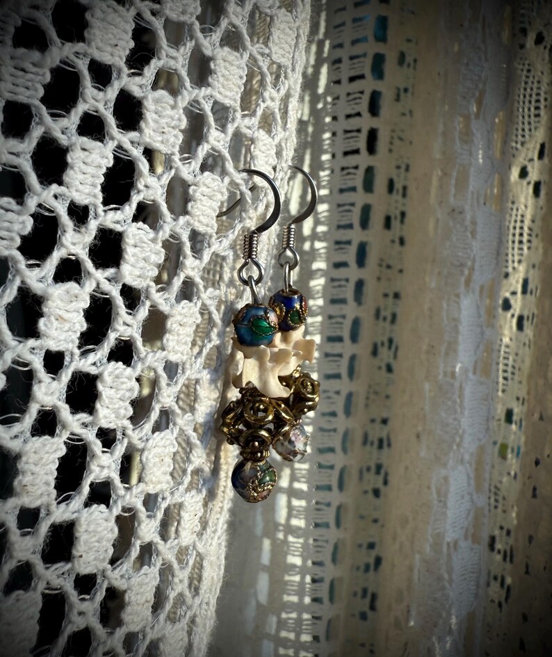 K&ouml;nnte beinhalten: Ein Paar Ohrringe mit einem wei&szlig;en knochenartigen Anh&auml;nger, blauen und gr&uuml;nen Emailleperlen und goldfarbenen Akzenten.