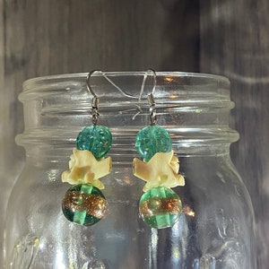 May include: A pair of dangle earrings with green and gold beads and a white bone-like charm. The earrings are hanging from a glass jar.
