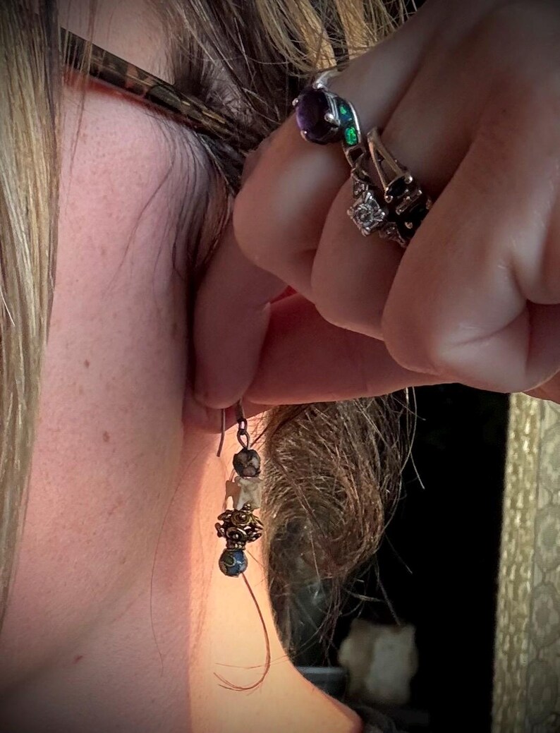 K&ouml;nnte beinhalten: Ein Ohrh&auml;nger mit einem blauen und goldenen Perlen, einer wei&szlig;en Knochenperle und einer braunen Perle. Der Ohrring h&auml;ngt am Ohr einer Person.