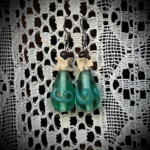 May include: A pair of handcrafted earrings featuring teal teardrop-shaped glass beads with a turquoise swirl design. The earrings have a bone-like accent and a dark brown bead at the top, suspended from silver-colored ear wires.