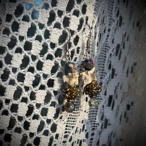 K&ouml;nnte beinhalten: Ein Paar Ohrringe mit einem Knochenanh&auml;nger, goldenen Perlen und bunten Emailleperlen. Die Ohrringe h&auml;ngen an silbernen Haken.