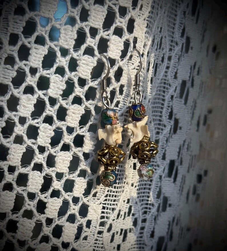 K&ouml;nnte beinhalten: Ein Paar Ohrringe mit einem Knochenanh&auml;nger, goldenen Perlen und bunten Emailleperlen. Die Ohrringe h&auml;ngen an silbernen Haken.