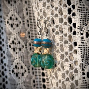 May include: Teal and gold glass bead earrings with a white swirl design. The earrings are hanging from silver hooks.