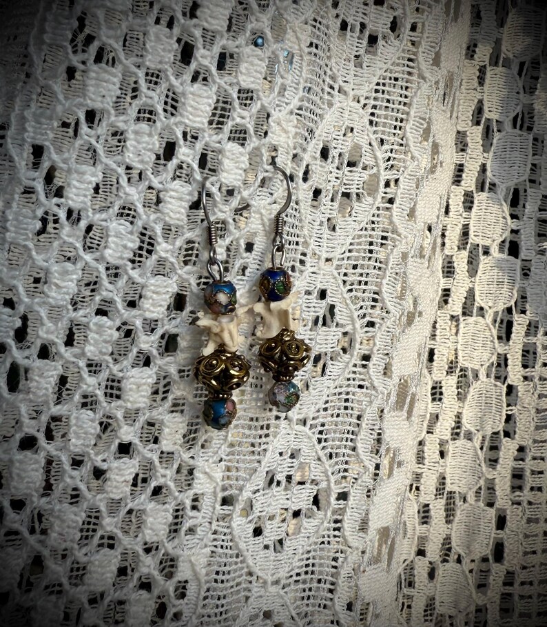 K&ouml;nnte beinhalten: Ein Paar Ohrringe mit wei&szlig;en knochenartigen Anh&auml;ngern, goldenen Perlen und blauen und wei&szlig;en gemusterten Glasperlen.