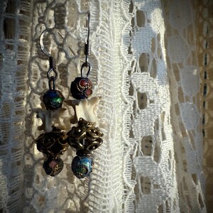 K&ouml;nnte beinhalten: Ein Paar H&auml;ngeohrringe mit einem Knochen-Anh&auml;nger, goldenen Perlen und bunten Emailleperlen. Die Ohrringe h&auml;ngen an einem wei&szlig;en Spitzenhintergrund.