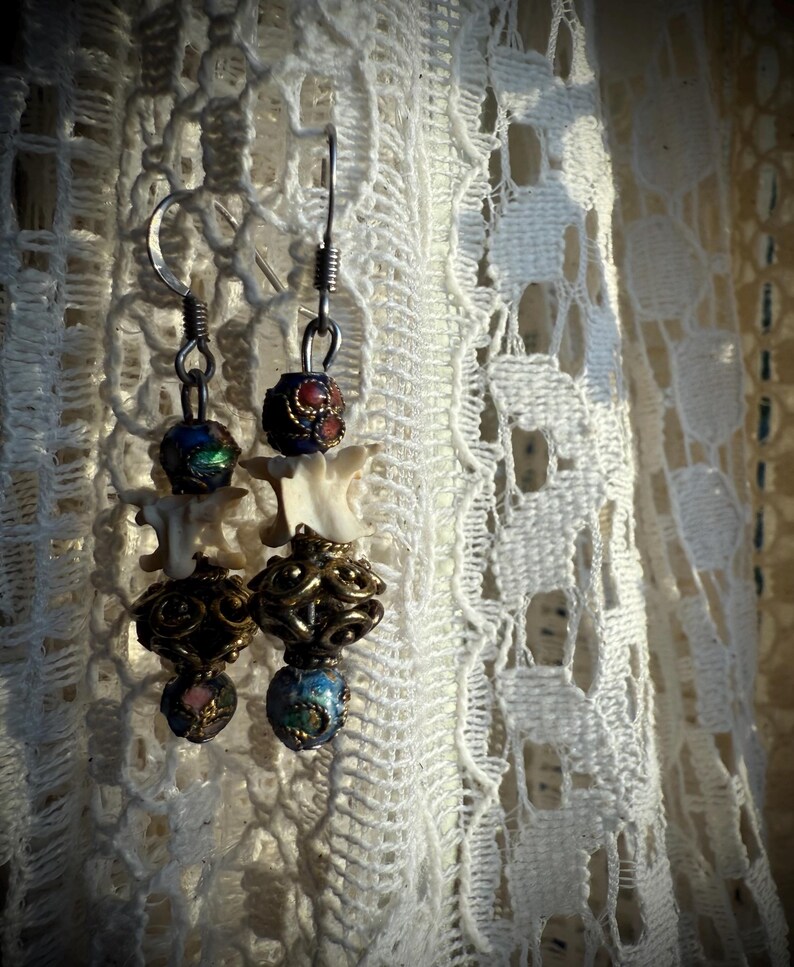 K&ouml;nnte beinhalten: Ein Paar H&auml;ngeohrringe mit einem Knochen-Anh&auml;nger, goldenen Perlen und bunten Emailleperlen. Die Ohrringe h&auml;ngen an einem wei&szlig;en Spitzenhintergrund.