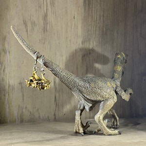 Puede incluir: Una figurita de dinosaurio marrón y gris con un pendiente dorado colgando de su cuello. El dinosaurio está de pie sobre una superficie de madera.