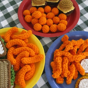 Pode incluir: Um close-up de itens de comida de crochê em pratos. Há sanduíches, batatas fritas, nuggets de frango e fatias de pizza de crochê. Os pratos são amarelos, vermelhos e azuis.