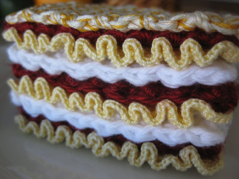 May include: A crocheted cake made with white, red, and yellow yarn. The cake is layered and has a scalloped edge.
