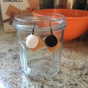 May include: Crochet earrings in a s'mores design. Each earring has a square, tan-coloured graham cracker base, topped with either a white marshmallow or a dark chocolate circle. The earrings hang from bronze-coloured ear wires.