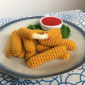 May include: A crocheted plate of food featuring golden-yellow mozzarella sticks, a small white bowl of red sauce, and green lettuce. One mozzarella stick is cut open, revealing a white interior. The plate has a blue rim.