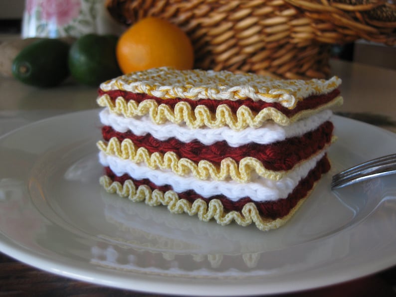 May include: A crocheted lasagna dish made with white, yellow, and red yarn. The dish is sitting on a white plate.