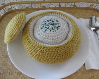 May include: A crocheted bowl with a lid, both in a yellow color. The bowl is filled with a white crocheted object with green embroidery. The bowl is sitting on a white plate.