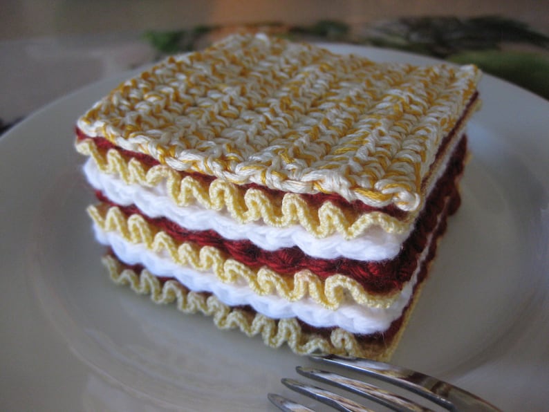 May include: A crocheted lasagna dish made with yellow, white, and red yarn. The dish is layered and has a realistic appearance.