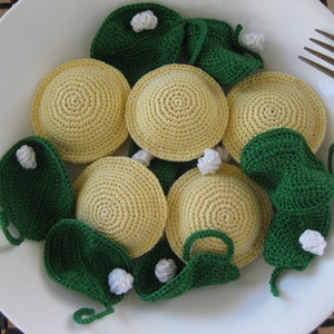 May include: A white plate holds a crocheted pasta dish. The dish features yellow ravioli and green spinach-like leaves, all with white accents. A fork rests on the plate's edge, suggesting a meal. The handmade food items are arranged to resemble a meal.