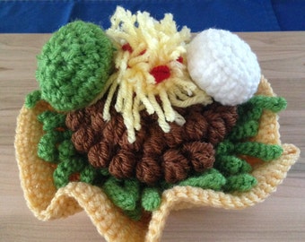 May include: A crocheted taco shell filled with brown meat, green lettuce, yellow cheese, and a white sour cream dollop.