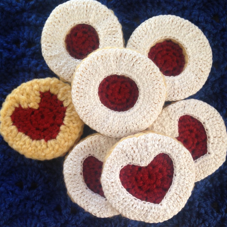 May include: Handmade crochet coasters in the shape of cookies. The coasters feature a cream-colored outer ring with a red center, some with a heart shape. The coasters are arranged on a blue surface.