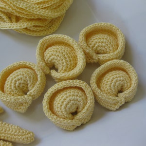 May include: A close-up of five crocheted yellow pasta shapes, resembling tortellini, on a white surface. There are also several crocheted yellow strands of pasta.