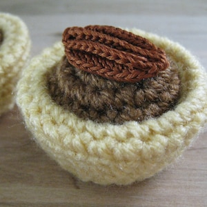 Puede incluir: Dos tartas de pastel de nuez pecan crochetadas con un relleno marrón y una nuez pecan en la parte superior. Las tartas están hechas con un hilo amarillo claro.