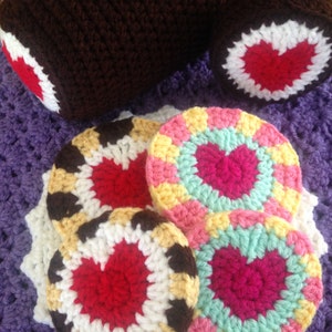 May include: Crocheted items in shades of brown, red, white, yellow, pink, and teal. The image features a brown crocheted log with red and white accents, along with several round coasters with heart designs.