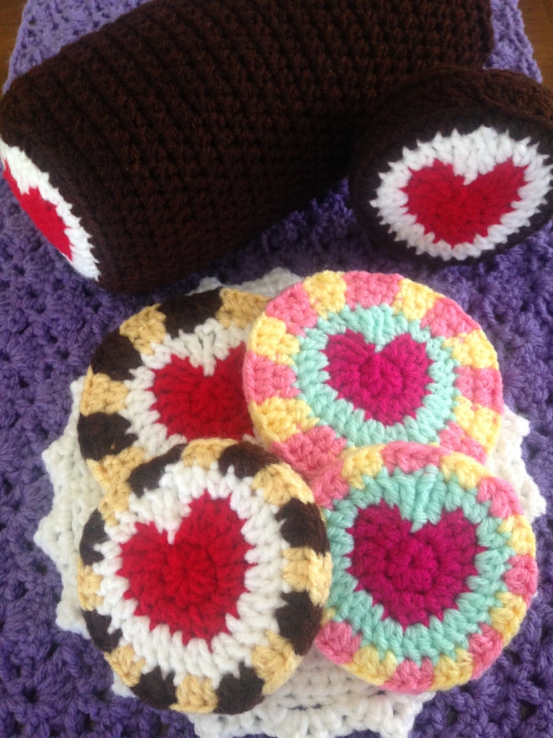May include: Crocheted items in shades of brown, red, white, yellow, pink, and teal. The image features a brown crocheted log with red and white accents, along with several round coasters with heart designs.