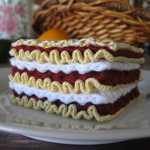 May include: A crocheted lasagna dish made with red, white, and yellow yarn. The dish is layered and has a ruffled edge.