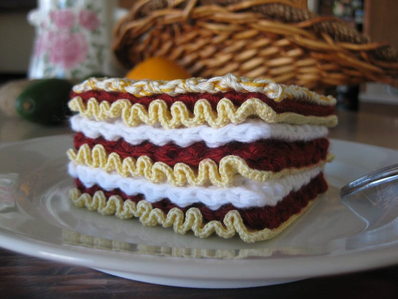 May include: A crocheted lasagna dish made with red, white, and yellow yarn. The dish is layered and has a ruffled edge.