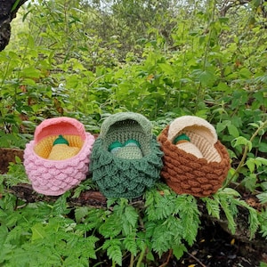 Könnte beinhalten: Drei gehäkelte Baby-Nester, eines rosa, eines grün und eines braun, sind auf einem Ast in einer Waldlandschaft angeordnet.