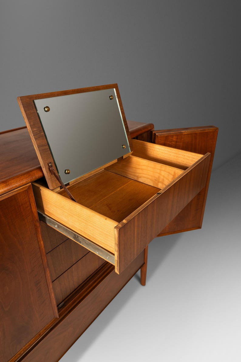 May include: A mid-century modern walnut dresser with a mirrored top and three drawers. The dresser has a warm brown finish and is in good condition.