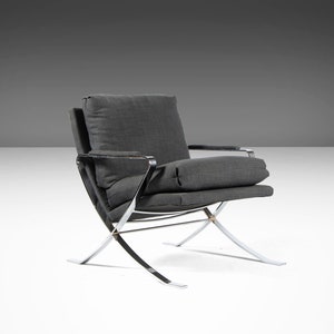May include: A gray upholstered armchair with a chrome frame. The chair has a simple design with a low back and a wide seat. The chair is on a white surface.