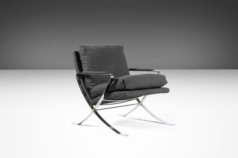 May include: A gray upholstered armchair with a chrome frame. The chair has a simple design with a low back and a wide seat. The chair is on a white surface.