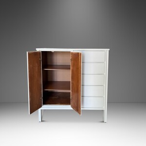 May include: A white cabinet with two doors open, revealing shelves. The cabinet has a series of drawers on the right side. The doors and interior shelves are a warm brown color, contrasting with the white exterior.