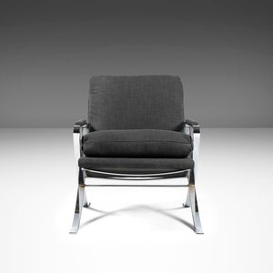 May include: A gray upholstered armchair with a chrome frame. The chair has a curved back and a wide seat. The chair is on a white surface.