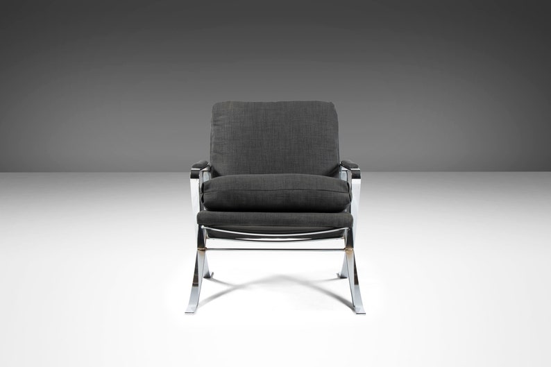 May include: A gray upholstered armchair with a chrome frame. The chair has a curved back and a wide seat. The chair is on a white surface.