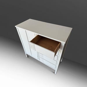 May include: A white wooden cabinet with a drawer open, revealing a brown interior. The cabinet has a rectangular shape with decorative geometric cutouts. The cabinet is set against a gray background.