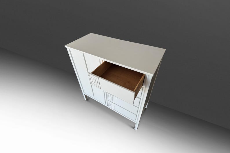 May include: A white wooden cabinet with a drawer open, revealing a brown interior. The cabinet has a rectangular shape with decorative geometric cutouts. The cabinet is set against a gray background.
