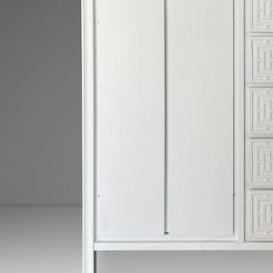 May include: A white wooden cabinet with a minimalist design. The cabinet features two doors and decorative geometric patterns on the right side. The cabinet is painted white and has a clean, modern aesthetic.