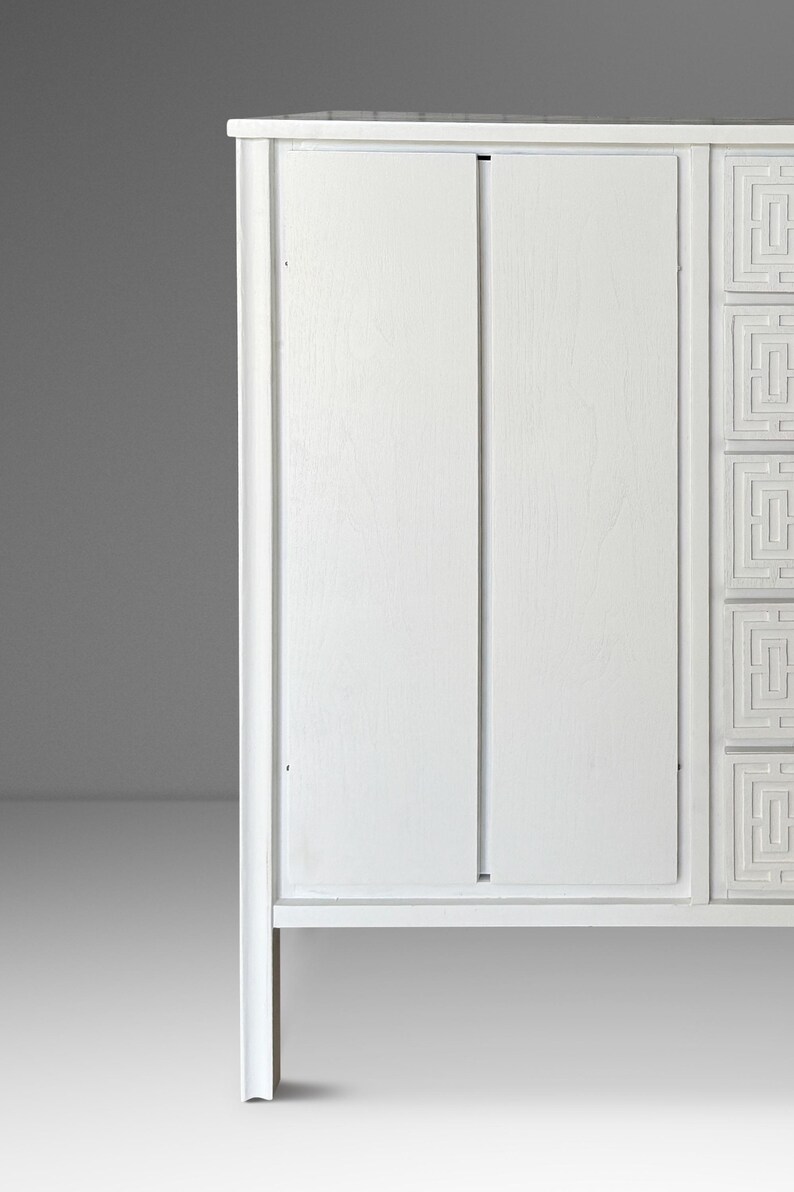 May include: A white wooden cabinet with a minimalist design. The cabinet features two doors and decorative geometric patterns on the right side. The cabinet is painted white and has a clean, modern aesthetic.